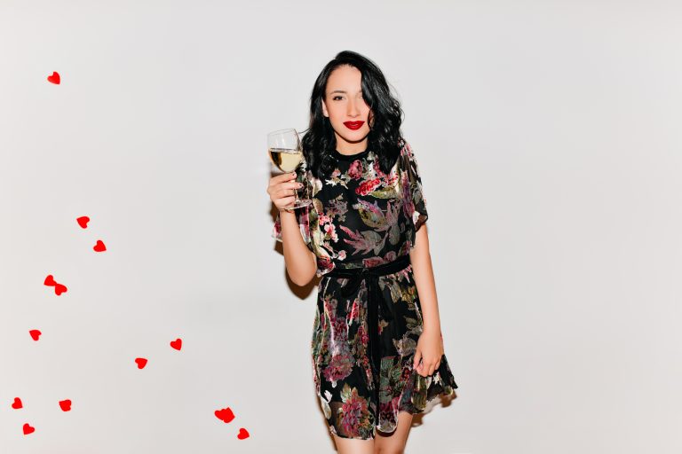 Lovely girl with black curly hair holding glass of champagne. Indoor portrait of enthusiastic european female model in dark dress drinking wine in valentine's day.